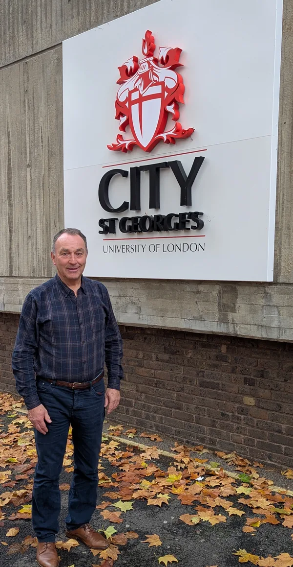Huw Edwards standing beneath the City St George's, University of London sign, with autumn leaves on the ground.