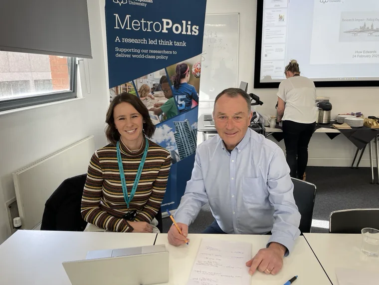Huw Edwards working one-to-one with a researcher at a desk, beside a banner for Metropolis, a research-led think tank.