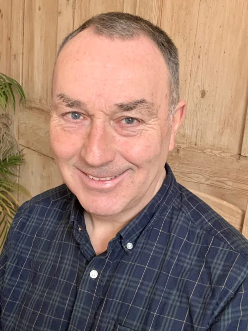 Portrait of Huw Edwards, smiling, in a dark checked shirt against a wooden wall.